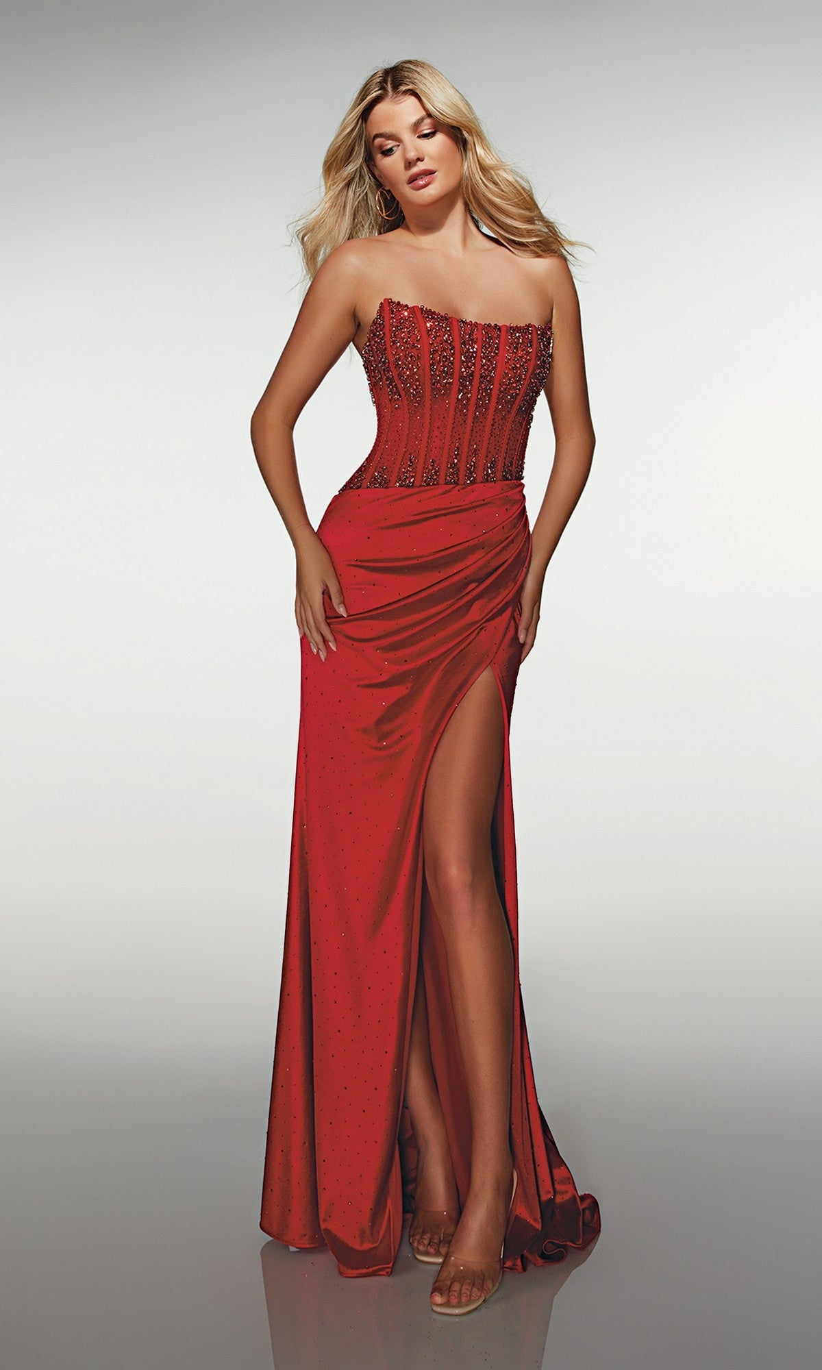 A woman poses in the Alyce 62147 long formal dress in red, featuring a strapless beaded corset bodice, high slit, and draped skirt, against a plain gradient background.